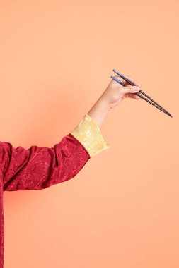Yeni Çin yılın kutlu olsun. Kırmızı geleneksel Cheongsam Qipao ya da Changshan elbisesi giyen Asyalı son sınıf öğrencisinin eli. Çin usulü yemek çubukları turuncu arka planda izole edilmiş..