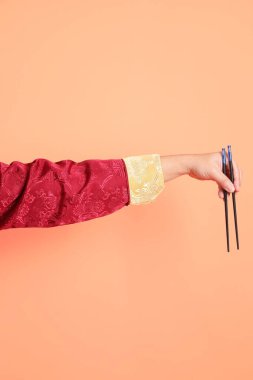 Yeni Çin yılın kutlu olsun. Kırmızı geleneksel Cheongsam Qipao ya da Changshan elbisesi giyen Asyalı son sınıf öğrencisinin eli. Çin usulü yemek çubukları turuncu arka planda izole edilmiş..