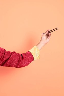 Yeni Çin yılın kutlu olsun. Kırmızı geleneksel Cheongsam Qipao ya da Changshan elbisesi giyen Asyalı son sınıf öğrencisinin eli. Çin usulü yemek çubukları turuncu arka planda izole edilmiş..