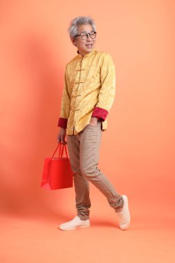 Yeni Çin yılın kutlu olsun. Asya 'lı enerjik son sınıf öğrencisi. Geleneksel altın kıyafetler giyiyor. El ele tutuşup turuncu arka planda izole edilmiş kağıt alışveriş torbaları taşıyor..