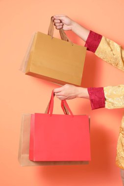 Yeni Çin yılın kutlu olsun. Turuncu arka planda izole edilmiş kağıt poşetlerle el ele tutuşan, geleneksel altın elbiseler giyen Asyalı son sınıf öğrencisinin eli..