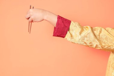 Yeni Çin yılın kutlu olsun. Asya 'lı son sınıf öğrencisi altın rengi Cheongsam Qipao ya da Changshan elbisesi giyiyor. Elinde gümüş çubuklarla turuncu arka planda duruyor..