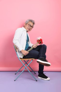 Pembe arka planda izole bir çiçek buketi tutarken sandalyede oturma jesti yapan Asyalı son sınıf öğrencisi. Sevgililer Günü