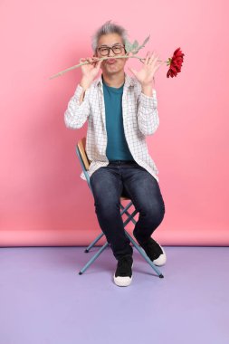 Pembe arka planda izole bir çiçek buketi tutarken sandalyede oturma jesti yapan Asyalı son sınıf öğrencisi. Sevgililer Günü