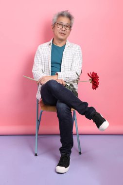 Pembe arka planda izole bir çiçek buketi tutarken sandalyede oturma jesti yapan Asyalı son sınıf öğrencisi. Sevgililer Günü