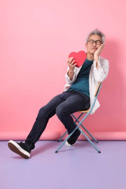 Asyalı üst düzey bir adam, pembe arka planda kırmızı kalp şeklinde izole bir şekilde otururken sandalyeye oturma jesti yapıyor. Sevgililer Günü