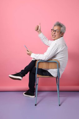 Pembe arka planda cep telefonunu kapalı tutarken sandalyeye oturma jesti yapan Asyalı son sınıf öğrencisi. Sevgililer Günü