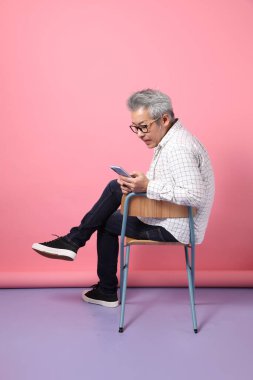 Pembe arka planda cep telefonunu kapalı tutarken sandalyeye oturma jesti yapan Asyalı son sınıf öğrencisi. Sevgililer Günü