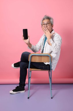 Pembe arka planda cep telefonunu kapalı tutarken sandalyeye oturma jesti yapan Asyalı son sınıf öğrencisi. Sevgililer Günü