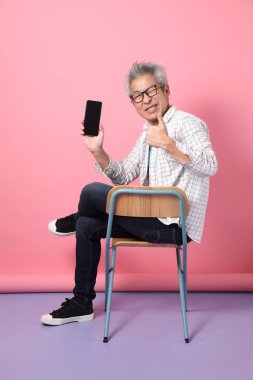 Pembe arka planda cep telefonunu kapalı tutarken sandalyeye oturma jesti yapan Asyalı son sınıf öğrencisi. Sevgililer Günü