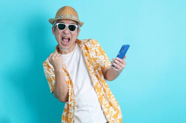 Elinde cep telefonuyla seyahat kıyafetleriyle mavi arka planda poz veren Asyalı enerjik kıdemli adamın portresi. Olgun adam, Asyalı adam. Yaşlılar, Tatil, Seyahat. Stüdyo. Çoklu duygular.