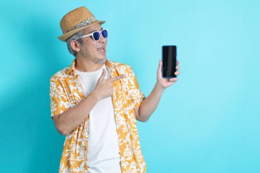 Elinde cep telefonuyla seyahat kıyafetleriyle mavi arka planda poz veren Asyalı enerjik kıdemli adamın portresi. Olgun adam, Asyalı adam. Yaşlılar, Tatil, Seyahat. Stüdyo. Çoklu duygular.