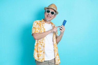 Mavi arka planda seyahat kıyafetleriyle izole edilmiş cep telefonu tutan Asyalı enerjik kıdemli adamın portresi. Olgun adam, Asyalı adam. Yaşlılar, Tatil, Seyahat. Stüdyo. Çoklu duygular.