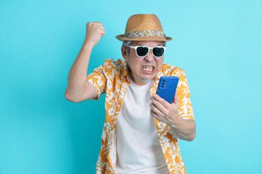 Mavi arka planda seyahat kıyafetleriyle izole edilmiş cep telefonu tutan Asyalı enerjik kıdemli adamın portresi. Olgun adam, Asyalı adam. Yaşlılar, Tatil, Seyahat. Stüdyo. Çoklu duygular.