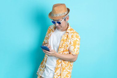 Mavi arka planda seyahat kıyafetleriyle izole edilmiş cep telefonu tutan Asyalı enerjik kıdemli adamın portresi. Olgun adam, Asyalı adam. Yaşlılar, Tatil, Seyahat. Stüdyo. Çoklu duygular.