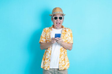 Mavi arka planda seyahat kıyafetleriyle izole edilmiş cep telefonu tutan Asyalı enerjik kıdemli adamın portresi. Olgun adam, Asyalı adam. Yaşlılar, Tatil, Seyahat. Stüdyo. Çoklu duygular.
