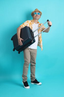 Mavi arka planda, elinde güneş gözlüğü olan Asyalı enerjik yaşlı bir adam bavulu tek başına tutuyor. Olgun adam, Asyalı adam. Yaşlı, stüdyo fotoğrafı. Çoklu duygular. Seyahat kavramı