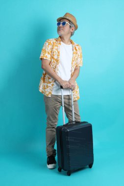 Mavi arka planda, elinde güneş gözlüğü olan Asyalı enerjik yaşlı bir adam bavulu tek başına tutuyor. Olgun adam, Asyalı adam. Yaşlı, stüdyo fotoğrafı. Çoklu duygular. Seyahat kavramı