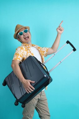 Mavi arka planda, elinde güneş gözlüğü olan Asyalı enerjik yaşlı bir adam bavulu tek başına tutuyor. Olgun adam, Asyalı adam. Yaşlı, stüdyo fotoğrafı. Çoklu duygular. Seyahat kavramı