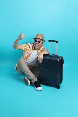 Mavi arka planda, elinde güneş gözlüğü olan Asyalı enerjik yaşlı bir adam bavulu tek başına tutuyor. Olgun adam, Asyalı adam. Yaşlı, stüdyo fotoğrafı. Çoklu duygular. Seyahat kavramı