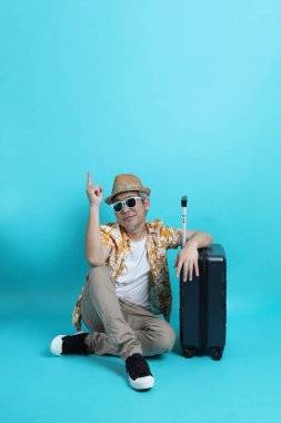 Mavi arka planda, elinde güneş gözlüğü olan Asyalı enerjik yaşlı bir adam bavulu tek başına tutuyor. Olgun adam, Asyalı adam. Yaşlı, stüdyo fotoğrafı. Çoklu duygular. Seyahat kavramı
