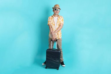Mavi arka planda, elinde güneş gözlüğü olan Asyalı enerjik yaşlı bir adam bavulu tek başına tutuyor. Olgun adam, Asyalı adam. Yaşlı, stüdyo fotoğrafı. Çoklu duygular. Seyahat kavramı