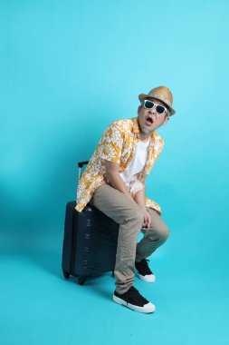 Mavi arka planda, elinde güneş gözlüğü olan Asyalı enerjik yaşlı bir adam bavulu tek başına tutuyor. Olgun adam, Asyalı adam. Yaşlı, stüdyo fotoğrafı. Çoklu duygular. Seyahat kavramı