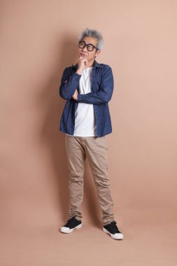 Mavi arka planda seyahat kıyafetleriyle izole edilmiş cep telefonu tutan Asyalı enerjik kıdemli adamın portresi. Olgun adam, Asyalı adam. Yaşlılar, Tatil, Seyahat. Stüdyo. Çoklu duygular.