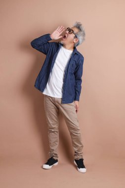 Mavi arka planda seyahat kıyafetleriyle izole edilmiş cep telefonu tutan Asyalı enerjik kıdemli adamın portresi. Olgun adam, Asyalı adam. Yaşlılar, Tatil, Seyahat. Stüdyo. Çoklu duygular.