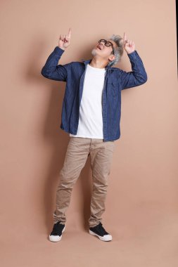 Mavi arka planda seyahat kıyafetleriyle izole edilmiş cep telefonu tutan Asyalı enerjik kıdemli adamın portresi. Olgun adam, Asyalı adam. Yaşlılar, Tatil, Seyahat. Stüdyo. Çoklu duygular.