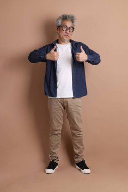 Mavi arka planda seyahat kıyafetleriyle izole edilmiş cep telefonu tutan Asyalı enerjik kıdemli adamın portresi. Olgun adam, Asyalı adam. Yaşlılar, Tatil, Seyahat. Stüdyo. Çoklu duygular.