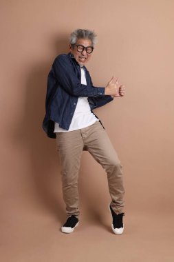 Mavi arka planda seyahat kıyafetleriyle izole edilmiş cep telefonu tutan Asyalı enerjik kıdemli adamın portresi. Olgun adam, Asyalı adam. Yaşlılar, Tatil, Seyahat. Stüdyo. Çoklu duygular.