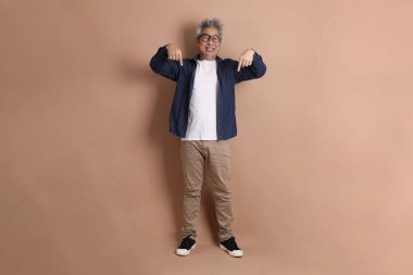 Mavi arka planda seyahat kıyafetleriyle izole edilmiş cep telefonu tutan Asyalı enerjik kıdemli adamın portresi. Olgun adam, Asyalı adam. Yaşlılar, Tatil, Seyahat. Stüdyo. Çoklu duygular.