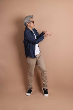 Mavi arka planda seyahat kıyafetleriyle izole edilmiş cep telefonu tutan Asyalı enerjik kıdemli adamın portresi. Olgun adam, Asyalı adam. Yaşlılar, Tatil, Seyahat. Stüdyo. Çoklu duygular.