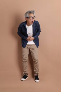 Gözlüklü, Asyalı, enerjik, üst düzey bir adam kahverengi arka planda izole edilmiş ve günlük kıyafetlerle poz veriyor. Olgun adam, Asyalı adam. Yaşlı, stüdyo fotoğrafı. Çoklu duygular.