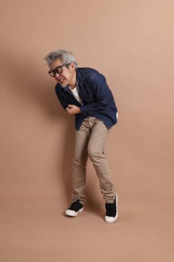 Gözlüklü, Asyalı, enerjik, üst düzey bir adam kahverengi arka planda izole edilmiş ve günlük kıyafetlerle poz veriyor. Olgun adam, Asyalı adam. Yaşlı, stüdyo fotoğrafı. Çoklu duygular.