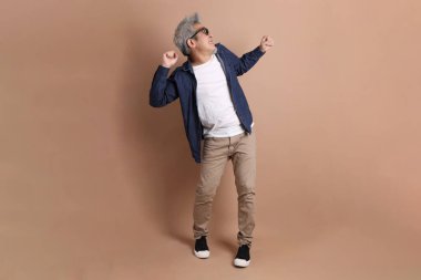 Gözlüklü, Asyalı, enerjik, üst düzey bir adam kahverengi arka planda izole edilmiş ve günlük kıyafetlerle poz veriyor. Olgun adam, Asyalı adam. Yaşlı, stüdyo fotoğrafı. Çoklu duygular.
