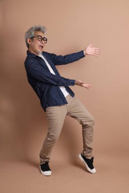 Gözlüklü, Asyalı, enerjik, üst düzey bir adam kahverengi arka planda izole edilmiş ve günlük kıyafetlerle poz veriyor. Olgun adam, Asyalı adam. Yaşlı, stüdyo fotoğrafı. Çoklu duygular.