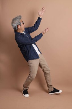Gözlüklü, Asyalı, enerjik, üst düzey bir adam kahverengi arka planda izole edilmiş ve günlük kıyafetlerle poz veriyor. Olgun adam, Asyalı adam. Yaşlı, stüdyo fotoğrafı. Çoklu duygular.