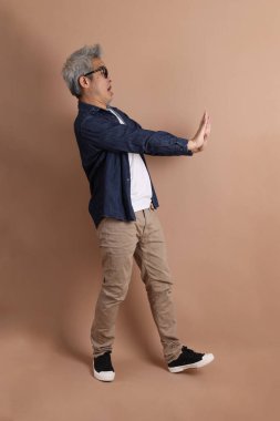 Gözlüklü, Asyalı, enerjik, üst düzey bir adam kahverengi arka planda izole edilmiş ve günlük kıyafetlerle poz veriyor. Olgun adam, Asyalı adam. Yaşlı, stüdyo fotoğrafı. Çoklu duygular.