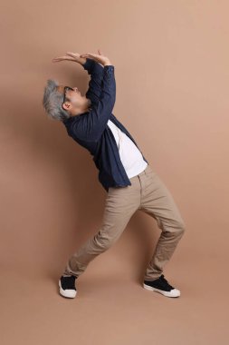 Gözlüklü, Asyalı, enerjik, üst düzey bir adam kahverengi arka planda izole edilmiş ve günlük kıyafetlerle poz veriyor. Olgun adam, Asyalı adam. Yaşlı, stüdyo fotoğrafı. Çoklu duygular.