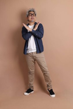 Gözlüklü, Asyalı, enerjik, üst düzey bir adam kahverengi arka planda izole edilmiş ve günlük kıyafetlerle poz veriyor. Olgun adam, Asyalı adam. Yaşlı, stüdyo fotoğrafı. Çoklu duygular.