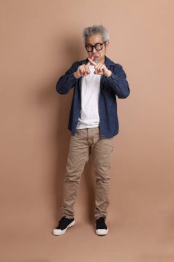 Gözlüklü, Asyalı, enerjik, üst düzey bir adam kahverengi arka planda izole edilmiş ve günlük kıyafetlerle poz veriyor. Olgun adam, Asyalı adam. Yaşlı, stüdyo fotoğrafı. Çoklu duygular.