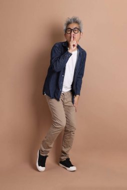 Gözlüklü, Asyalı, enerjik, üst düzey bir adam kahverengi arka planda izole edilmiş ve günlük kıyafetlerle poz veriyor. Olgun adam, Asyalı adam. Yaşlı, stüdyo fotoğrafı. Çoklu duygular.