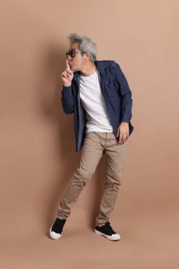 Gözlüklü, Asyalı, enerjik, üst düzey bir adam kahverengi arka planda izole edilmiş ve günlük kıyafetlerle poz veriyor. Olgun adam, Asyalı adam. Yaşlı, stüdyo fotoğrafı. Çoklu duygular.