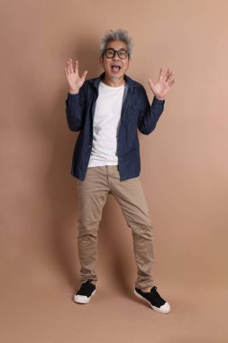 Gözlüklü, Asyalı, enerjik, üst düzey bir adam kahverengi arka planda izole edilmiş ve günlük kıyafetlerle poz veriyor. Olgun adam, Asyalı adam. Yaşlı, stüdyo fotoğrafı. Çoklu duygular.