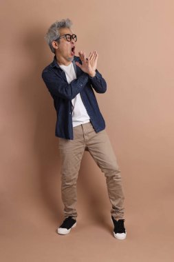 Gözlüklü, Asyalı, enerjik, üst düzey bir adam kahverengi arka planda izole edilmiş ve günlük kıyafetlerle poz veriyor. Olgun adam, Asyalı adam. Yaşlı, stüdyo fotoğrafı. Çoklu duygular.