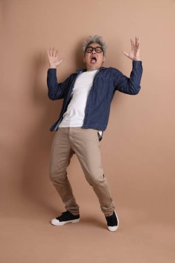 Gözlüklü, Asyalı, enerjik, üst düzey bir adam kahverengi arka planda izole edilmiş ve günlük kıyafetlerle poz veriyor. Olgun adam, Asyalı adam. Yaşlı, stüdyo fotoğrafı. Çoklu duygular.