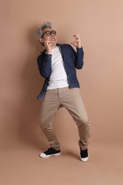 Gözlüklü, Asyalı, enerjik, üst düzey bir adam kahverengi arka planda izole edilmiş ve günlük kıyafetlerle poz veriyor. Olgun adam, Asyalı adam. Yaşlı, stüdyo fotoğrafı. Çoklu duygular.