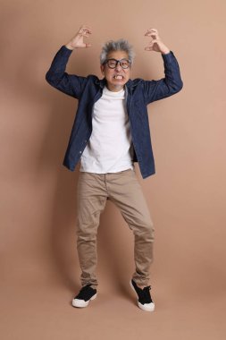 Gözlüklü, Asyalı, enerjik, üst düzey bir adam kahverengi arka planda izole edilmiş ve günlük kıyafetlerle poz veriyor. Olgun adam, Asyalı adam. Yaşlı, stüdyo fotoğrafı. Çoklu duygular.